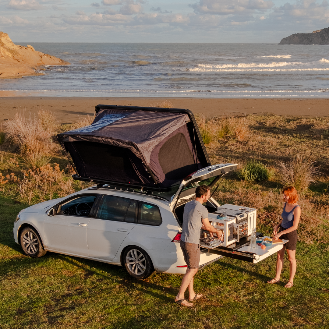 Stel kookt op het strand met campingbox in de kofferbak van de stationwagon en uitgeschoven uitschuifsysteem met een daktent bij zonneschijn