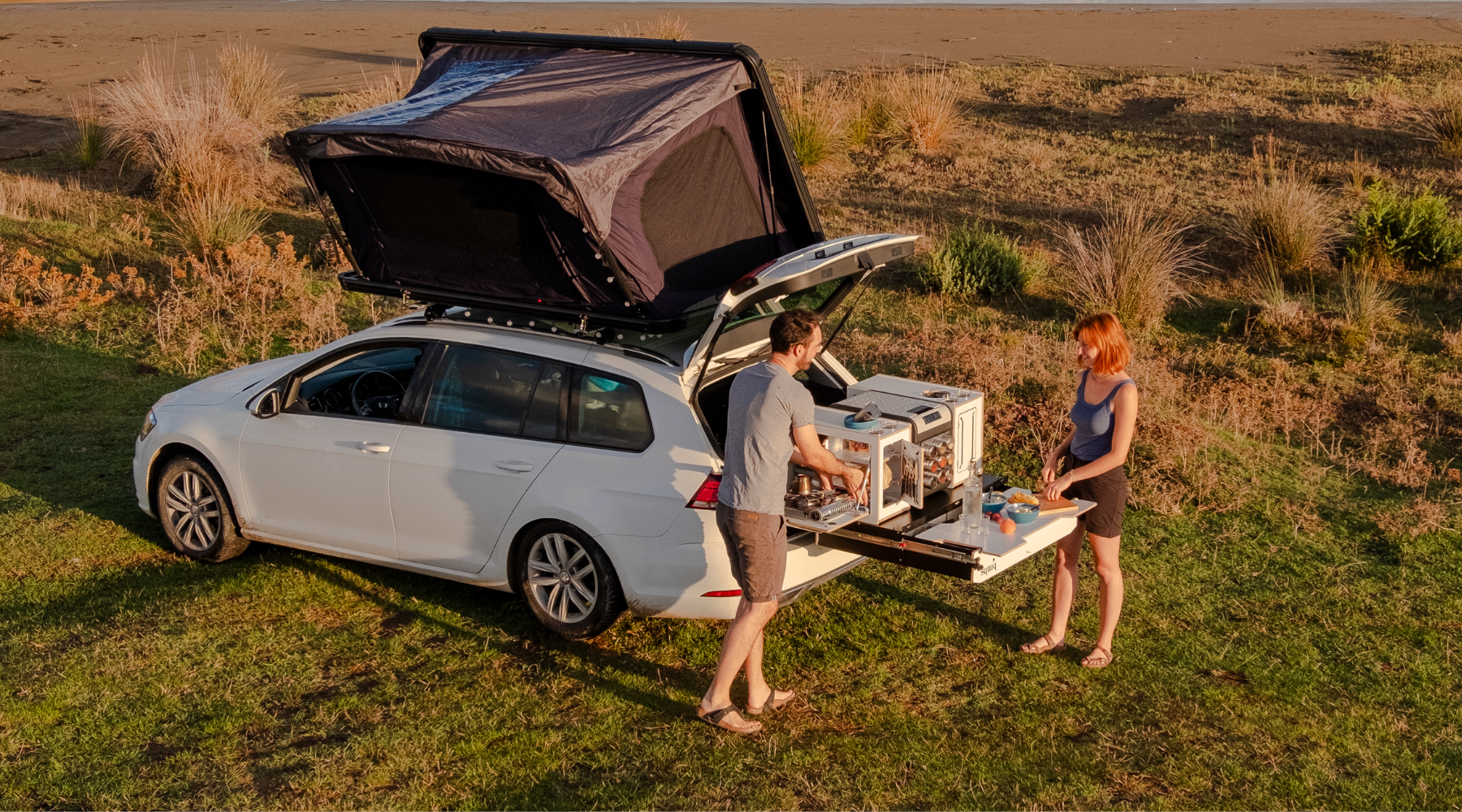 Paar kocht am Strand mit ausgezogenem Kofferraumauszug und Campingbox von SQUIQ – Auto mit Dachzelt als mobile Outdoor-Küche beim Camping