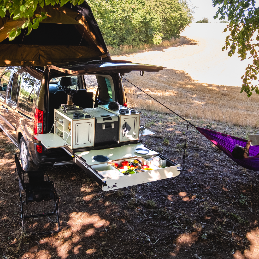 SQUIQ campingbox in de hoge dakcombi met daktent, hangmat en gedekte tafel, persoon leest ontspannen