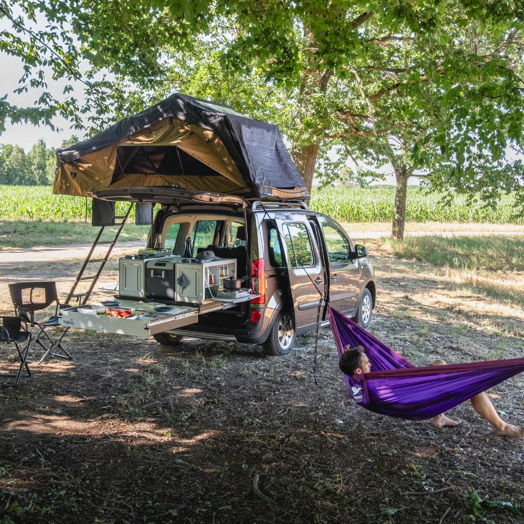 SQUIQ campingbox in de hoge dakcombi met daktent, gedekte tafel en persoon in hangmat in het groen