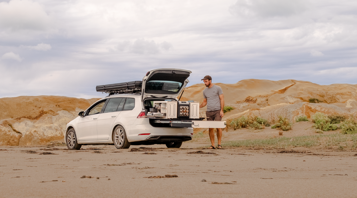 Break avec tiroir de coffre et tente de toit sur la plage, une personne se tient devant la campingbox.