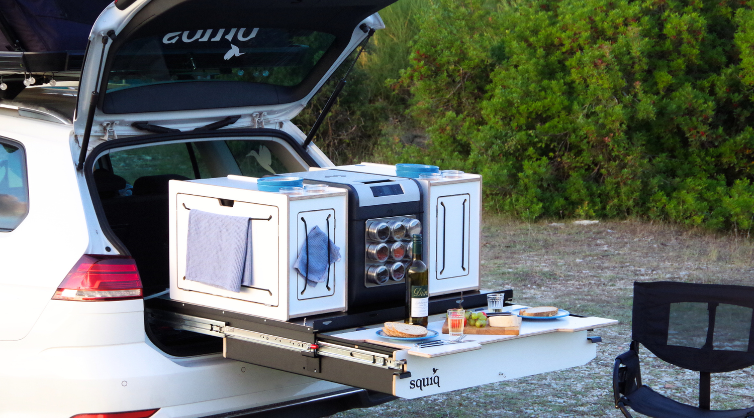 Ordnungssystem im Kofferraum für Camping mit Auto. Esstisch am Heck mit Campingstuhl und Essen.