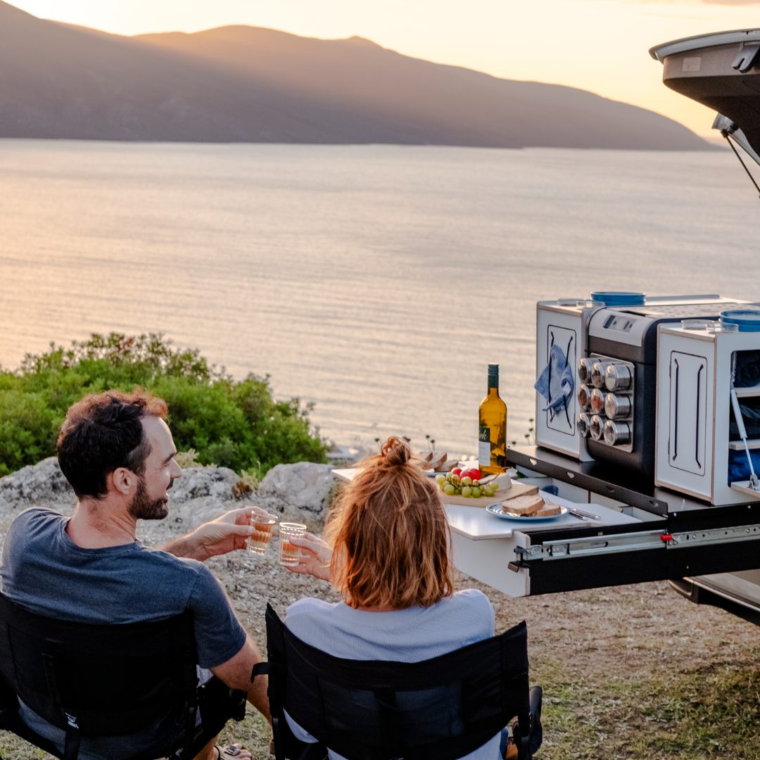 Zwei Personen am Mini-Camper-Ausbau mit Campingbox-Modulen und Esstisch mit Blick auf das Meer