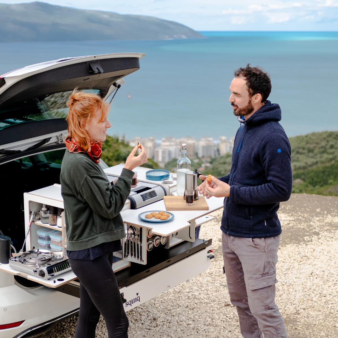 Twee personen eten bij een achterkeuken met campingboxen in een stationwagon, op de achtergrond de zee.