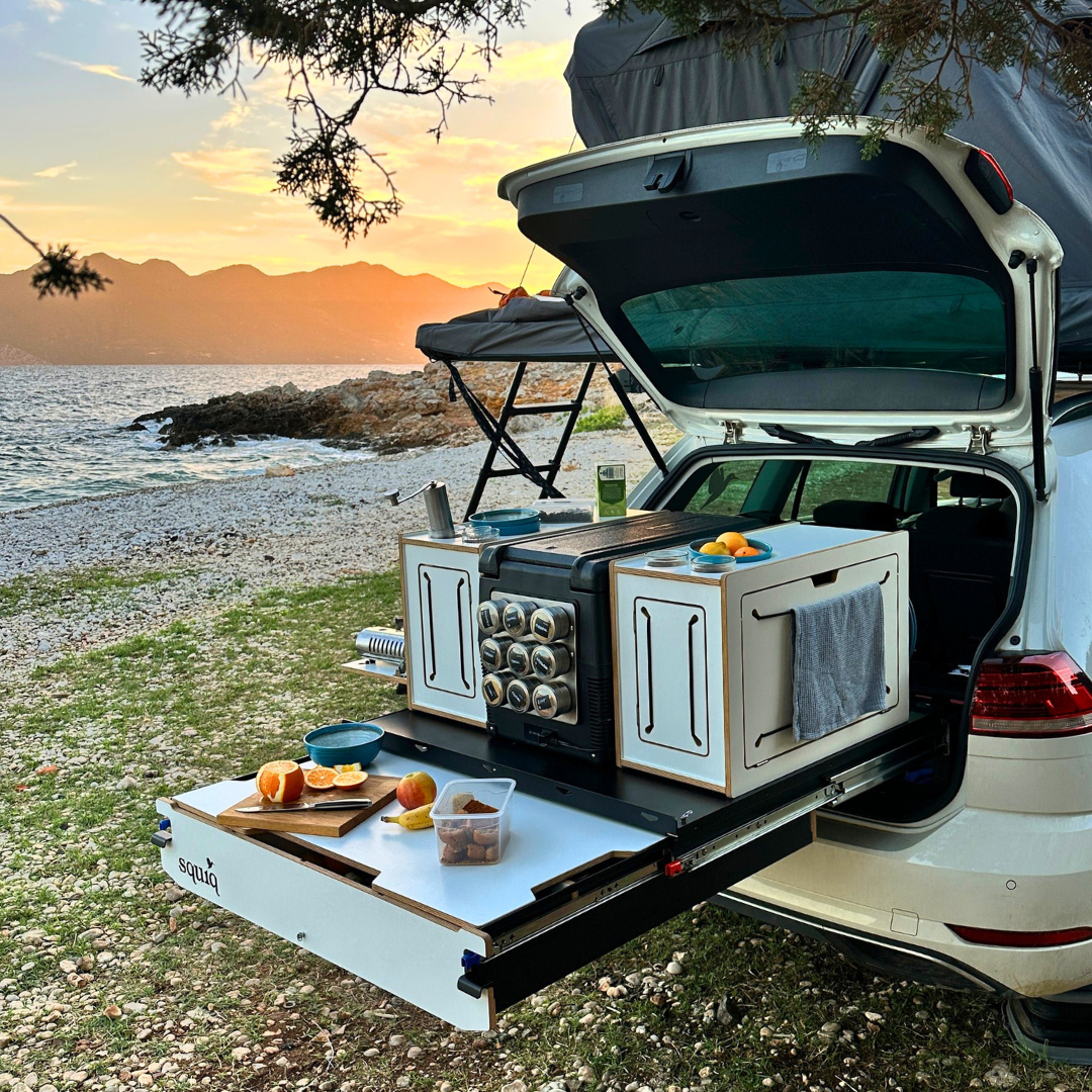 Campingbox in de kofferbak van een auto met daktent en uitgeschoven tafel tegen een zeekulisse tijdens het camperen met de auto