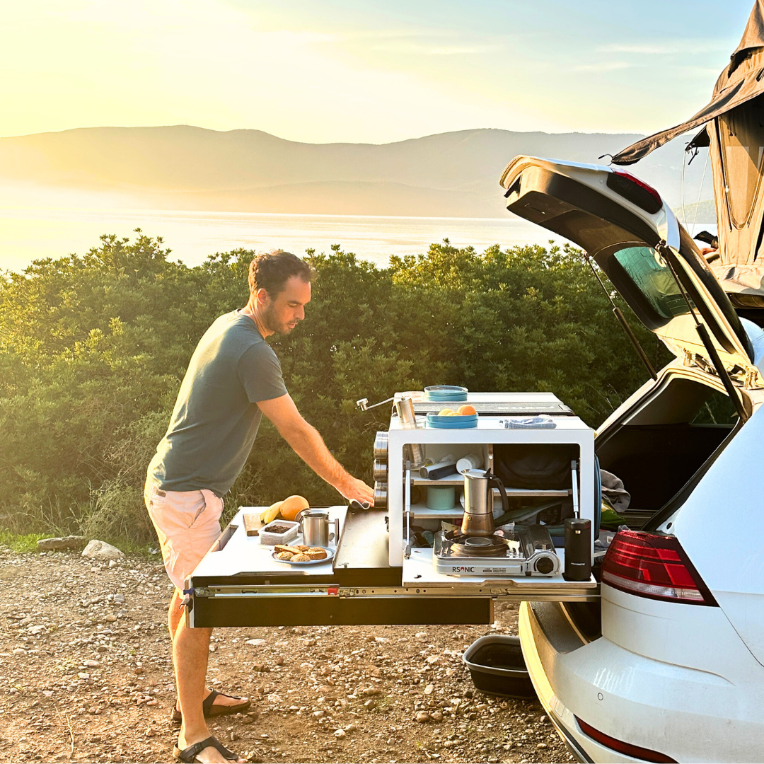 Persoon bereidt ontbijt bij een campingbox voor de auto in de kofferbak van een stationwagon met daktent, gaskooktoestel met koffie, zee en heuvels op de achtergrond