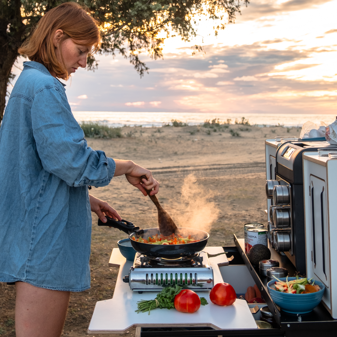 Person kocht an einer Camping Box mit Gaskocher und Pfanne, im Hintergrund der Strand und das Meer