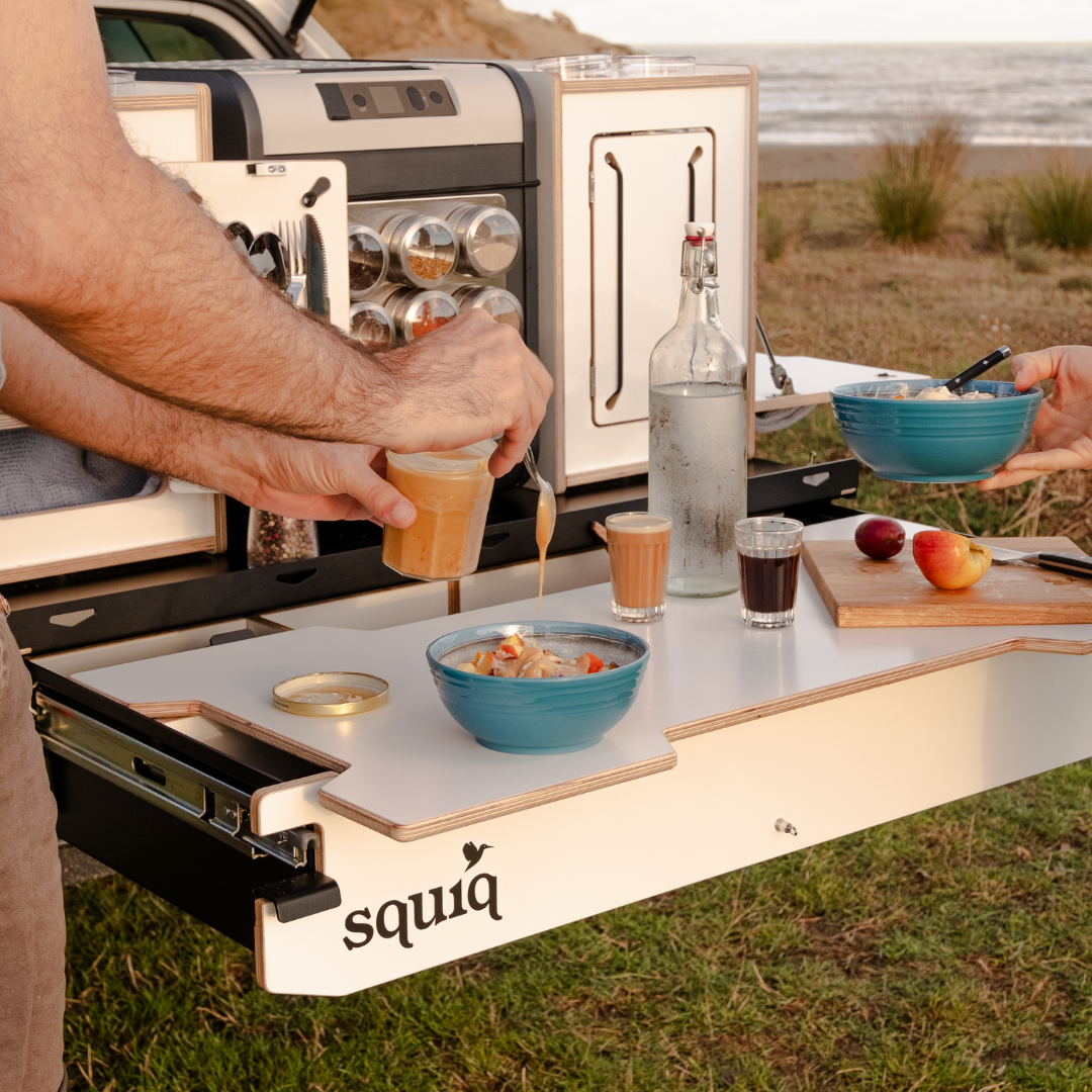 Ontbijt bereiden bij het SQUIQ uitschuifsysteem met campingbox in de kofferbak van de stationwagon en uitgeschoven uitschuifsysteem op het strand