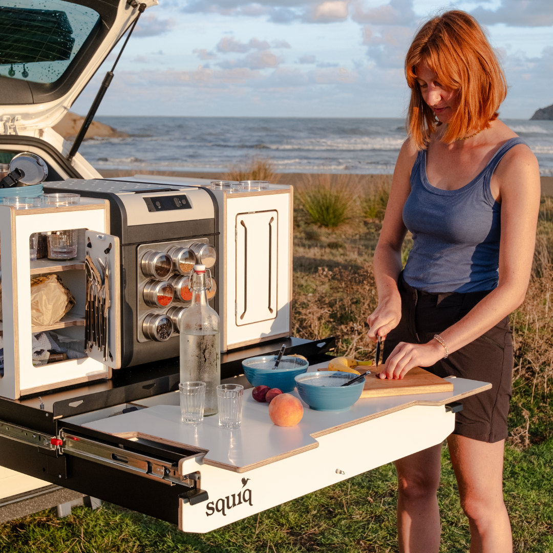 Vrouw snijdt fruit bij campingbox in de kofferbak van de stationwagon met uitgeschoven squiq uitschuifsysteem op het strand