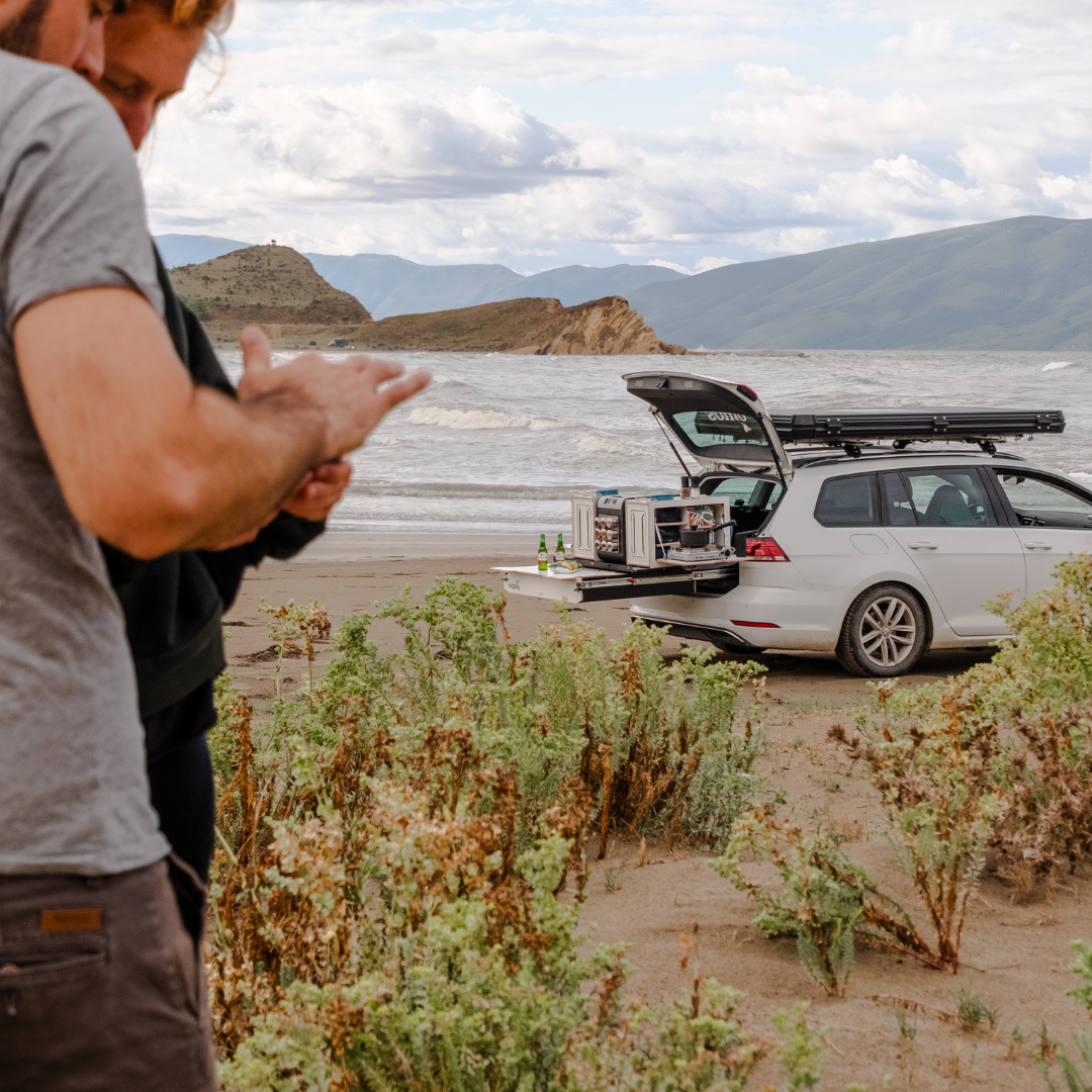 Stel kijkt op de telefoon voor een campingbox in de kofferbak van de stationwagon met uitgeschoven uitschuifsysteem aan zee