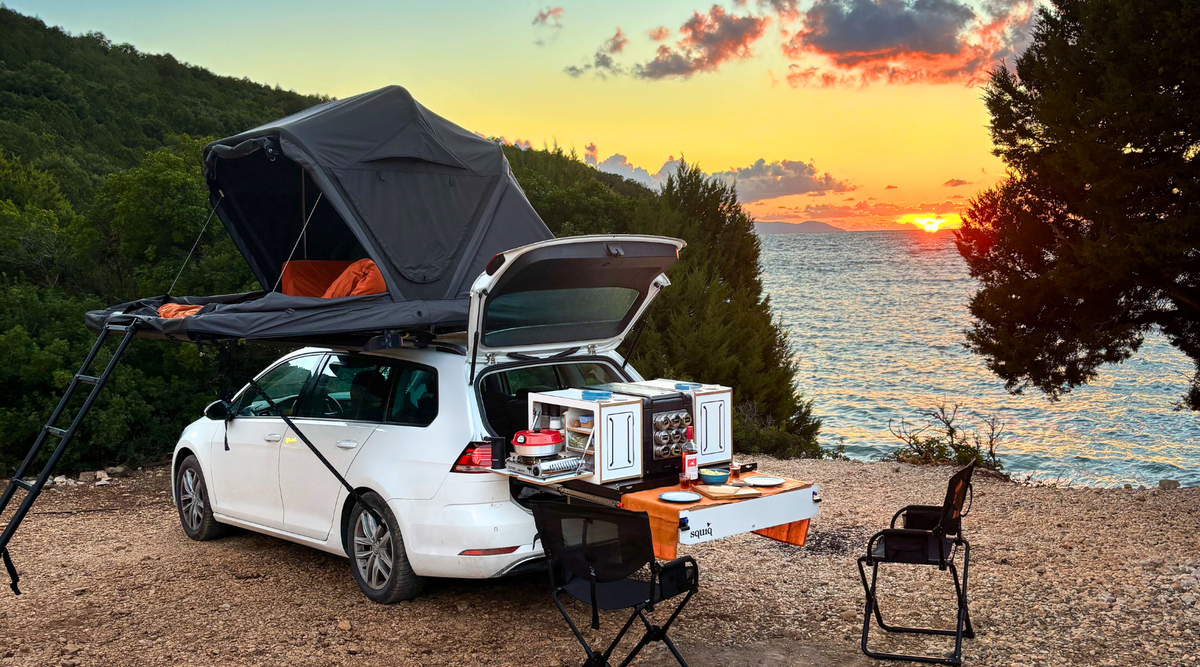 Campingbox in de kofferbak van een auto met eettafel en campingstoelen tijdens het kamperen met auto en daktent bij zonsondergang aan zee