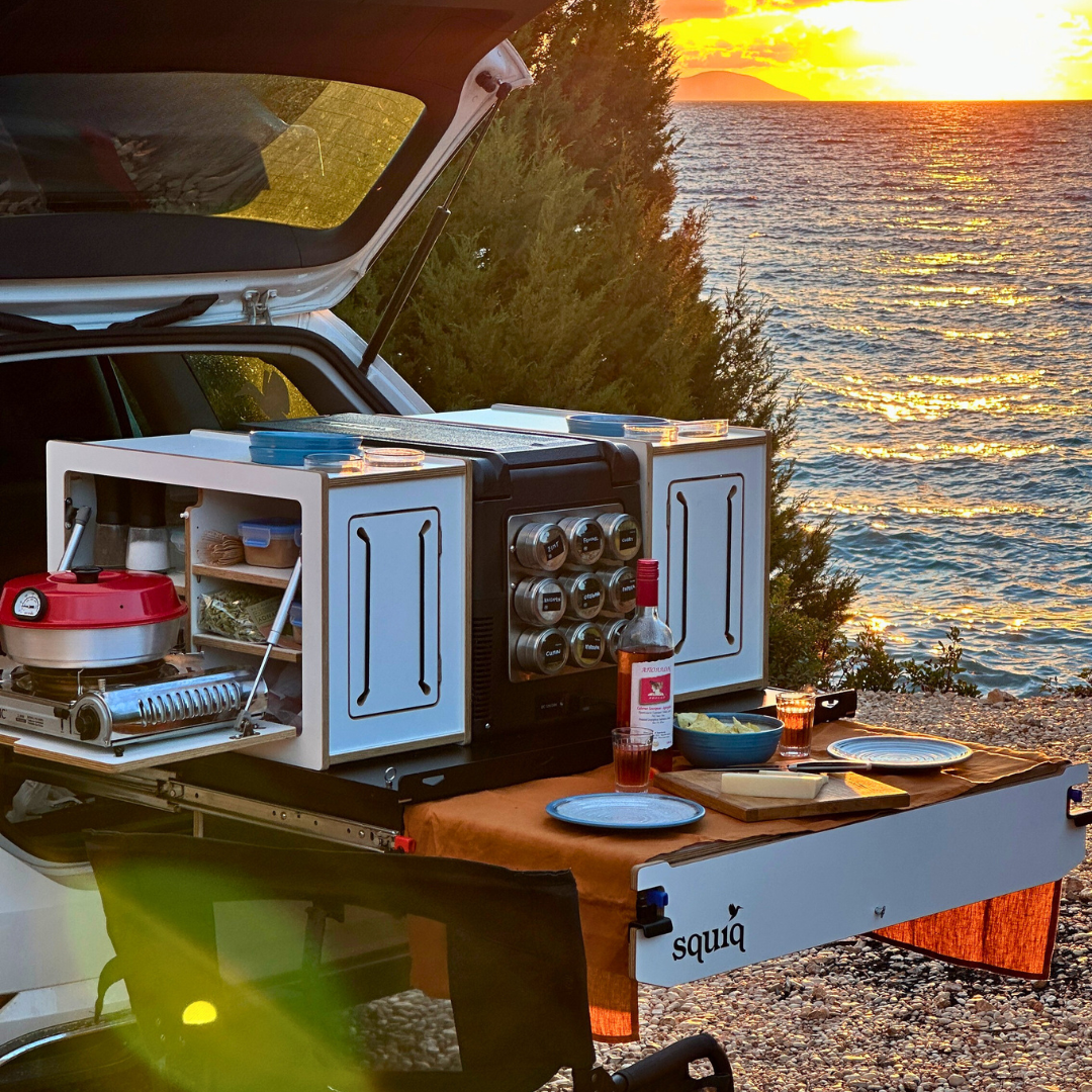 campingbox im Kofferraum von squiq mit Essen und Esstisch vor einer Strandkulisse bei Sonnenuntergang am Meer